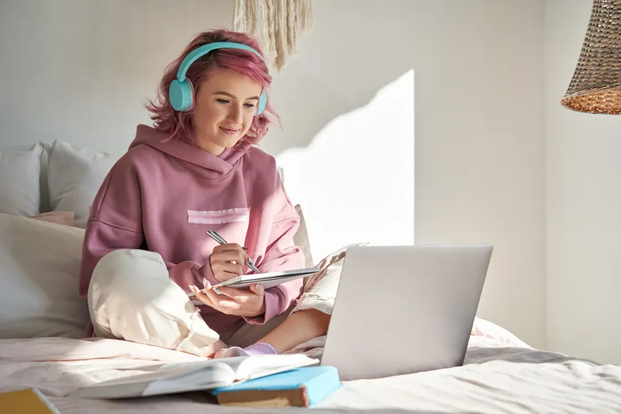 Bilde av en ung jente som sitter på en seng foran en pc og har på seg øretelefoner - illustrasjonsbilde